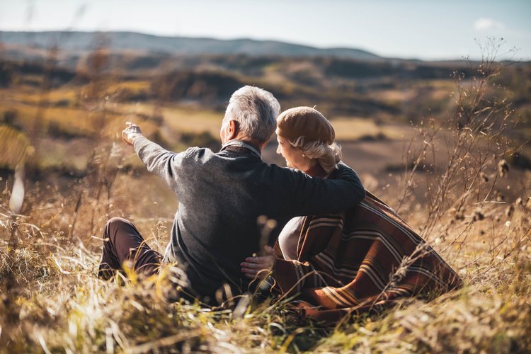 Back view of embraced senior couple relaxing in tall grass on a hill while man is aiming at distance.